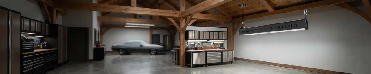 Spacious rustic-industrial garage workshop featuring exposed wooden beam ceilings and long suspended LED linear pendant lights illuminating a classic car and metal workbench.
