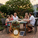 A family of five gathers around an outdoor dining table in a backyard patio setting, illuminated by warm string lights and a glowing solar-powered lantern. A small solar panel is visible on the brick ground next to the table.