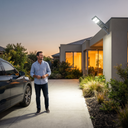 A man stands on a concrete driveway in front of a modern house at dusk, with a wall-mounted solar light illuminating the walkway and exterior. A dark SUV is partially visible on the left.