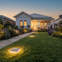 A landscaped front yard at dusk with a white house, lawn, flower beds, and warm ground lights. A square illuminated outdoor light is visible in the foreground grass.