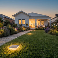 A landscaped front yard at dusk with a white house, lawn, flower beds, and warm ground lights. A square illuminated outdoor light is visible in the foreground grass.