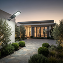 Modern single-story house exterior at dusk with a stone driveway, landscaped shrubs, olive trees, and a wall-mounted solar light fixture.