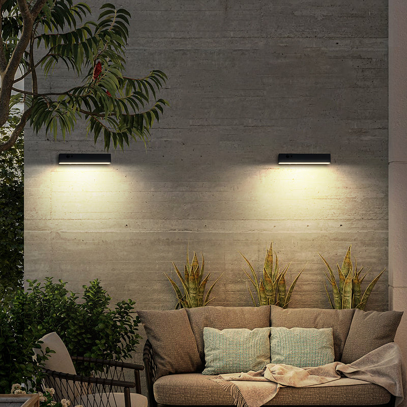 Two modern rectangular wall sconces mounted on a concrete patio wall, casting warm light downward. Below are outdoor plants and a beige sofa with pillows in a styled patio setting.