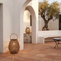 A woven natural-fiber lantern with a curved handle sits on terracotta tile flooring in a sunlit Mediterranean-style courtyard with white stucco walls, arches, a bench, and an olive tree.