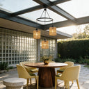 Three woven pendant lights hang over a round wooden dining table on a patio with cream chairs, glass block walls, and a glass roof in a sunlit outdoor setting.