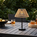 A modern table lamp with a black metal stand and a woven natural-fiber conical shade, placed on a dark outdoor patio table with drinks and greenery in the background.