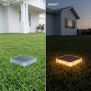 A split comparison showing a square solar ground mat on a green lawn. The left side is labeled 'DAY' and shows the mat as a cool, translucent gray-blue with no light emitted.