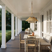 A woven wicker pendant light hangs over a dining table on a bright white porch with columns, chairs, and a cushioned bench.