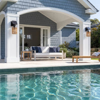 Outdoor patio seating area beside a swimming pool, featuring a white sofa with navy and white striped cushions, a wooden side table, a wooden bench, and a house with blue-gray shingles and white columns in the background.