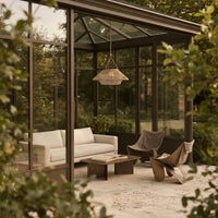 A modern outdoor patio area enclosed by a glass conservatory with dark metal framing, furnished with a white sofa, two sculptural brown chairs, a low wooden coffee table, and a woven pendant light. The scene is framed by soft-focus green foliage in the foreground.
