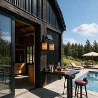 A modern outdoor bar setup with two black metal wall sconces featuring woven amber glass shades, mounted on a dark wood-clad building beside a pool deck with lounge chairs and a forest background.