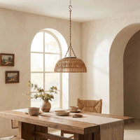 A woven wicker pendant light hangs over a rustic wooden dining table in a warm, neutral-toned dining room with wicker chairs and ceramic vessels.