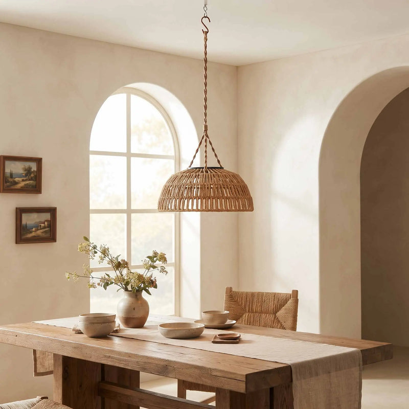 A woven wicker pendant light hangs over a rustic wooden dining table in a warm, neutral-toned dining room with wicker chairs and ceramic vessels.