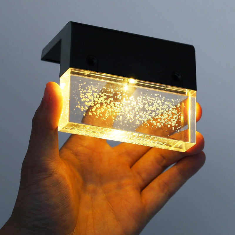 Hand holding a small rectangular light fixture with a black frame and clear glass cover against a gray background.