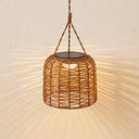 A hanging woven rattan pendant light with a warm glowing bulb inside, suspended by a rope from a ceiling, casting sunburst shadows on a textured beige wall.