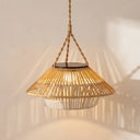 A natural bamboo and rattan pendant light with a woven shade, suspended by a thick rope from a solar panel, casting warm light on a textured beige wall.