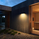 Wall-mounted outdoor light fixtures illuminating a concrete building facade at dusk, with a wooden door, deck, grass, and sky in the background.