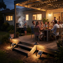 Outdoor patio scene at dusk with a group of people dining under warm string lights on a wooden deck beside a house. Solar ground lights and a small solar panel are visible near the steps, and one person is grilling on a barbecue grill with smoke rising.