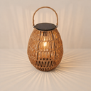 A large woven rattan lantern with a handle sits on terracotta tiles in a white arched courtyard, casting warm radial shadows near an olive tree and built-in bench.