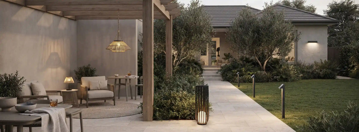 Modern outdoor patio and garden pathway at dusk, featuring a covered seating area with neutral-toned chairs and tables, a hanging pendant light, a tall black outdoor lantern, and a walkway leading to a house with illuminated walls and landscaping.