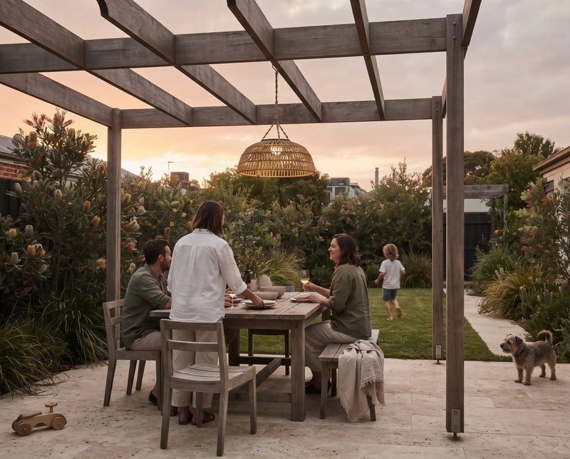 A family shares a meal at a wooden dining table under a wooden pergola in a landscaped backyard at sunset, with a hanging woven pendant light above and a small dog on the patio.