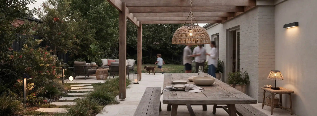 Outdoor patio scene under a wooden pergola with a large woven pendant light, a wooden dining table, and people walking in the background near a white brick house.