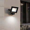 A black Outlux outdoor floodlight with a built-in motion sensor, mounted on a textured beige wall and casting a bright beam of light downward. The background shows a softly blurred modern building facade at night.