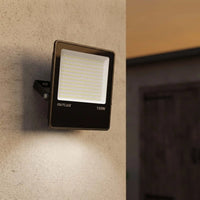 An OUTLUX 150W outdoor LED floodlight mounted on a textured beige wall, casting light downward, with a blurred interior doorway in the background.