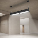 A modern rectangular industrial ceiling light fixture with a black housing and bright white illuminated panels, suspended from the ceiling in a minimalist concrete and plaster interior space.