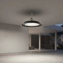 A modern circular pendant light with a black textured outer ring and a white illuminated center, suspended from a ceiling in a minimalist concrete pavilion.