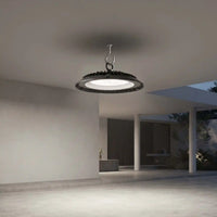A modern circular pendant light with a black textured outer ring and a white illuminated center, suspended from a ceiling in a minimalist concrete pavilion.