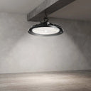 A large industrial-style LED light fixture with a black metal frame and a bright white diffuser, suspended from a concrete ceiling beam in a minimalist room with textured gray walls and wood-look flooring.