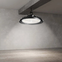 A large industrial-style LED light fixture with a black metal frame and a bright white diffuser, suspended from a concrete ceiling beam in a minimalist room with textured gray walls and wood-look flooring.