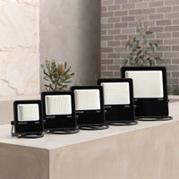 Four black outdoor LED floodlights of different wattages (30W, 50W, 70W, 150W) arranged in ascending size order on a concrete ledge, with a stone and brick wall background.