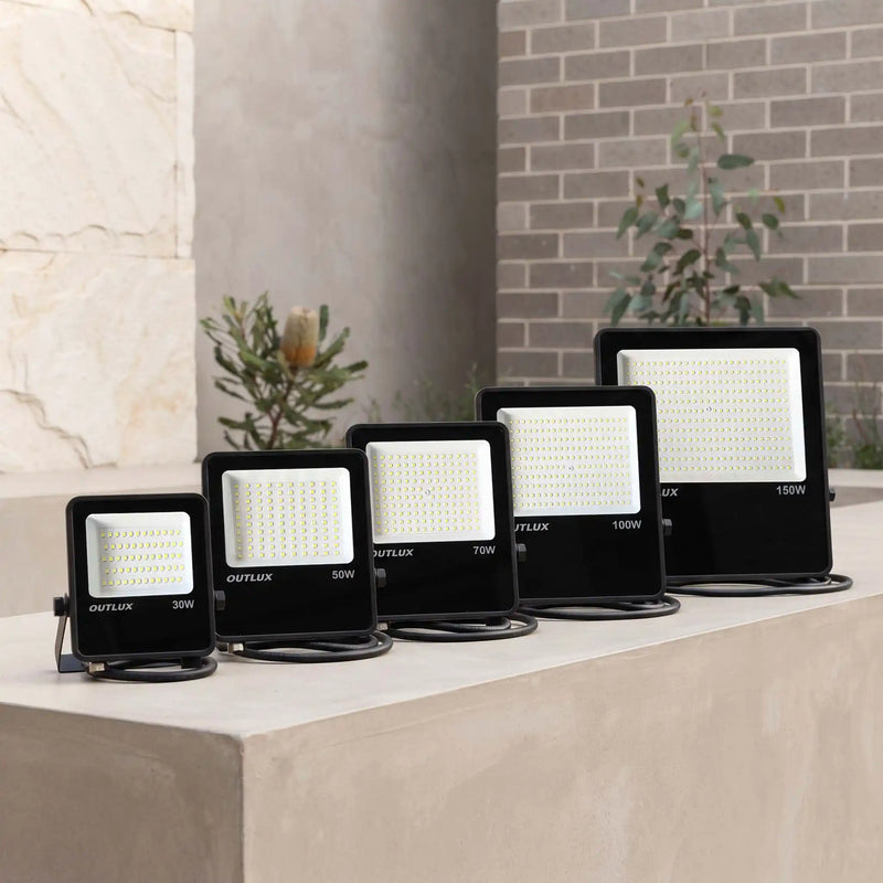 Four black outdoor LED floodlights of different wattages (30W, 50W, 70W, 150W) arranged in ascending size order on a concrete ledge, with a stone and brick wall background.