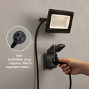 A black outdoor LED floodlight mounted on a gray wall, shown with a built-in SAA Australian wall outlet and a hand plugging in the cord. A circular inset highlights the three-pin Australian plug, with text noting an approximate 150cm exposed cable.